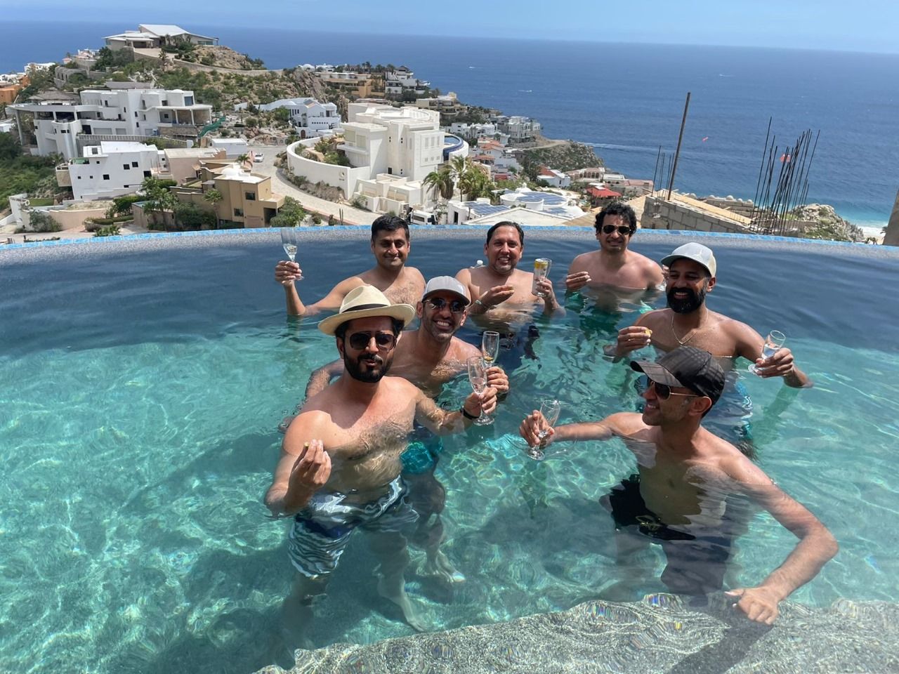 Seven men toasting with champagne glasses in a sunlit infinity pool overlooking white hillside villas and the blue ocean — ocean-view coastal celebration.