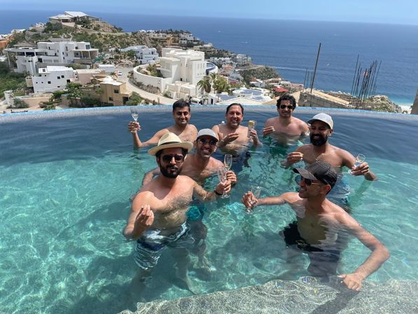 Seven men toasting with champagne glasses in a sunlit infinity pool overlooking white hillside villas and the blue ocean — ocean-view coastal celebration.