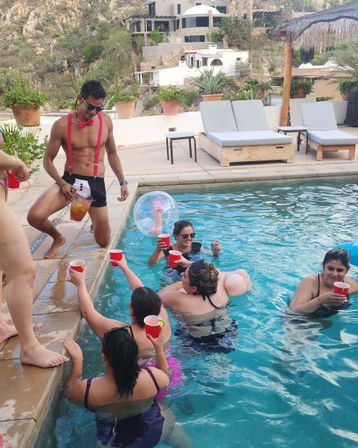 Lively pool party at a hillside villa — shirtless man in red suspenders and bow tie pours a pitcher while friends in swimsuits float, toast with red cups, and relax by sun loungers.