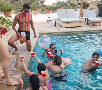 Lively pool party at a hillside villa — shirtless man in red suspenders and bow tie pours a pitcher while friends in swimsuits float, toast with red cups, and relax by sun loungers.