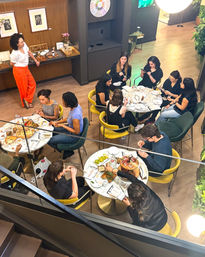 Overhead view of a lively modern cafe interior — groups gathered around round marble tables eating charcuterie boards and snacks, sipping drinks and chatting amid yellow and green chairs, wood floors, plants and a mezzanine staircase.