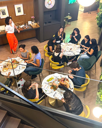Overhead view of a lively modern cafe interior — groups gathered around round marble tables eating charcuterie boards and snacks, sipping drinks and chatting amid yellow and green chairs, wood floors, plants and a mezzanine staircase.