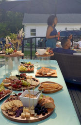 Backyard deck summer gathering with a colorful charcuterie spread on wooden boards—cheese, grapes, crackers, sliced baguette and nuts, with guests chatting under a patio umbrella.