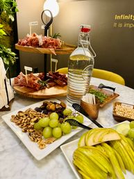 Two-tier wooden charcuterie stand with prosciutto and herbs, plates of green grapes, walnuts and dried figs, sliced pears, pine nuts, and a glass swing-top water bottle on a marble table — elegant appetizer spread.