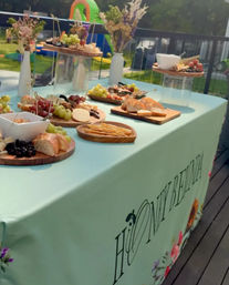 Backyard deck charcuterie spread on a mint tablecloth — wooden boards with bread, crackers, cheeses, grapes and dried fruit, decorative dried flower vases and a blurred kids’ inflatable playset on the lawn