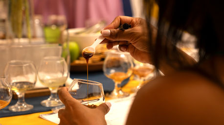 Close-up of hands drizzling dark syrup from a spoon into a snifter glass at an indoor spirits tasting table with multiple tasting glasses and warm ambient lighting.
