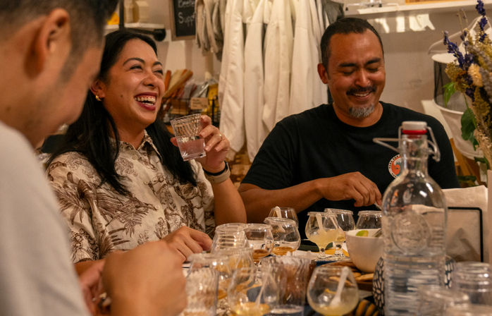 Group of people laughing at a cozy indoor tasting table, a woman holding a glass while others sample multiple small cocktail glasses, snacks and a large glass water bottle nearby.