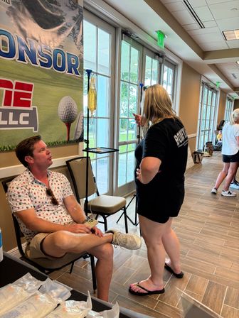 Indoor event lobby with glass doors, a seated man receiving an IV hydration drip from a hanging bag while chatting with a casually dressed woman in front of a golf-themed sponsor banner.
