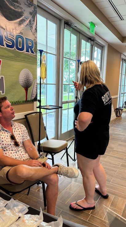Indoor event lobby with glass doors, a seated man receiving an IV hydration drip from a hanging bag while chatting with a casually dressed woman in front of a golf-themed sponsor banner.