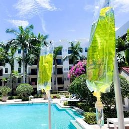 Poolside IV bags with yellow-green fluid hanging beside a sunny tropical resort pool framed by palm trees, bougainvillea, and a white multi-story courtyard building