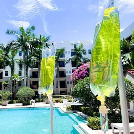 Poolside IV bags with yellow-green fluid hanging beside a sunny tropical resort pool framed by palm trees, bougainvillea, and a white multi-story courtyard building