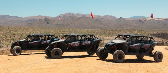 Three rugged off-road UTVs lined up on a sunbaked southwestern desert overlook, cacti-dotted valley and rocky mountain ridges under a clear blue sky with safety flags flying.