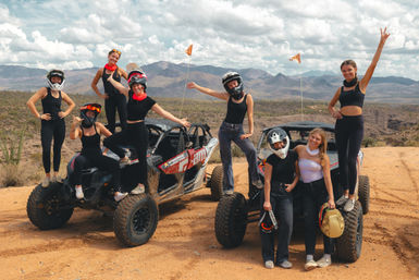 Group of friends posing on muddy side-by-side UTVs in a desert mountain landscape, wearing helmets and waving after an off-road adventure