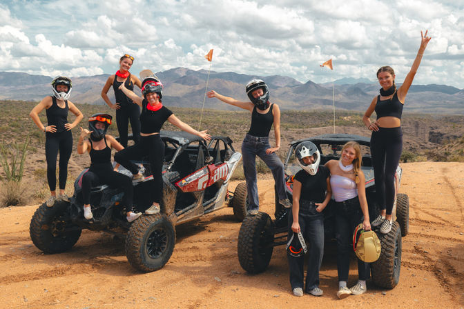 Group of friends posing on muddy side-by-side UTVs in a desert mountain landscape, wearing helmets and waving after an off-road adventure