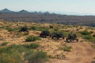 Four off-road UTVs kicking up dust on a sunlit dirt trail through arid desert scrub with layered mountain ridges in the background.
