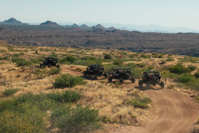 Four off-road UTVs kicking up dust on a sunlit dirt trail through arid desert scrub with layered mountain ridges in the background.