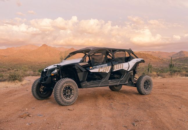 Four-seat off-road UTV parked on a dusty desert trail with tall cacti and rugged mountains under a golden sunset sky