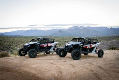 Two rugged four-seat off-road UTVs parked on a sandy desert overlook with scrub brush and a layered mountain range in the distance under a soft evening sky
