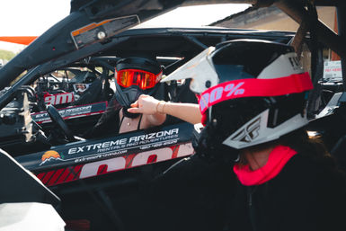 Two helmeted off-road riders in a side-by-side UTV at an Arizona desert rental, bright orange goggles and a pink helmet strap visible