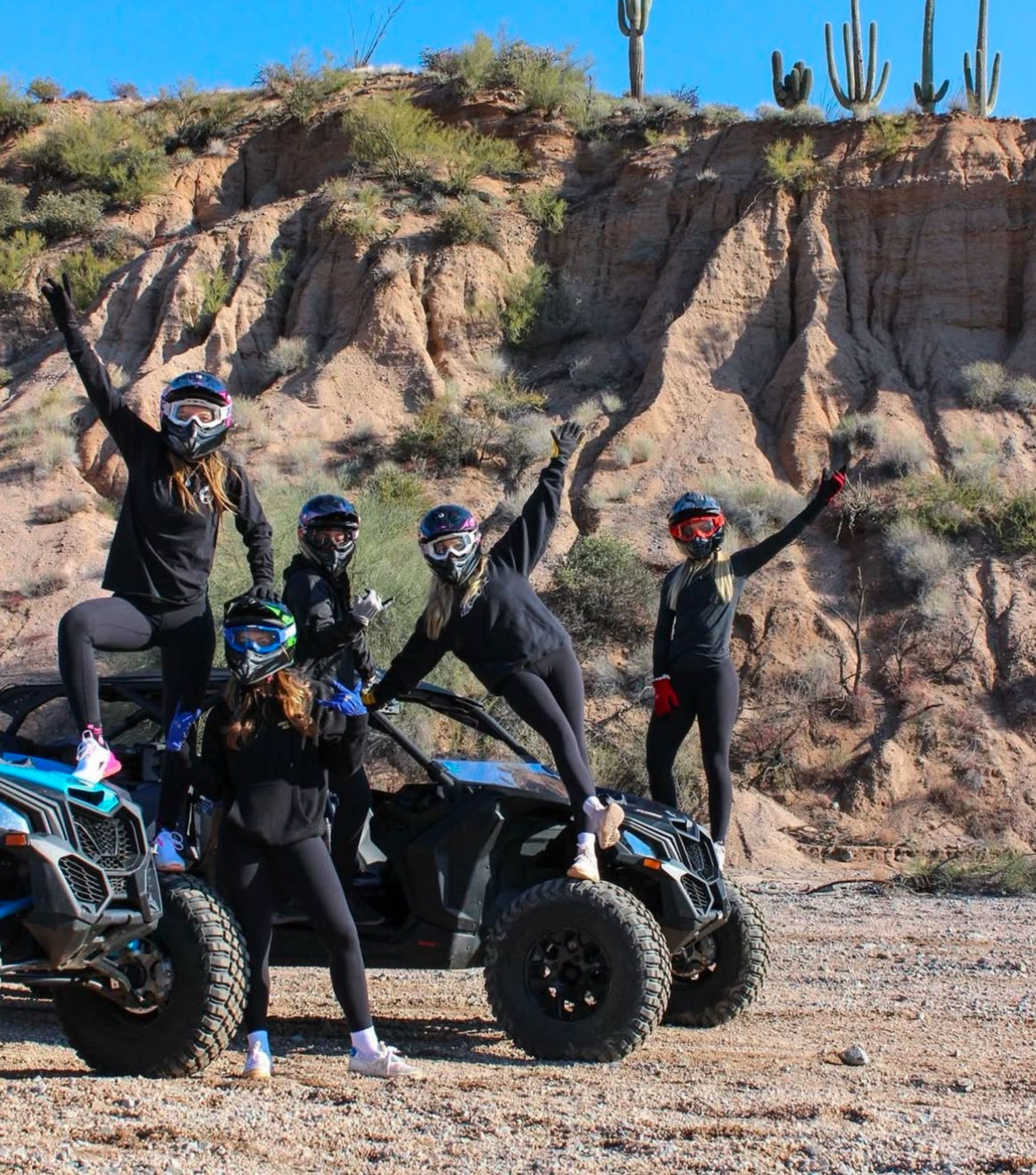 Five helmeted riders in black outfits posing on and beside two blue off-road UTVs in an Arizona desert, with saguaro cacti and red rock cliffs under a bright blue sky.