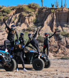 Five helmeted riders in black outfits posing on and beside two blue off-road UTVs in an Arizona desert, with saguaro cacti and red rock cliffs under a bright blue sky.