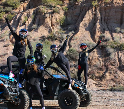 Five helmeted riders in black outfits posing on and beside two blue off-road UTVs in an Arizona desert, with saguaro cacti and red rock cliffs under a bright blue sky.