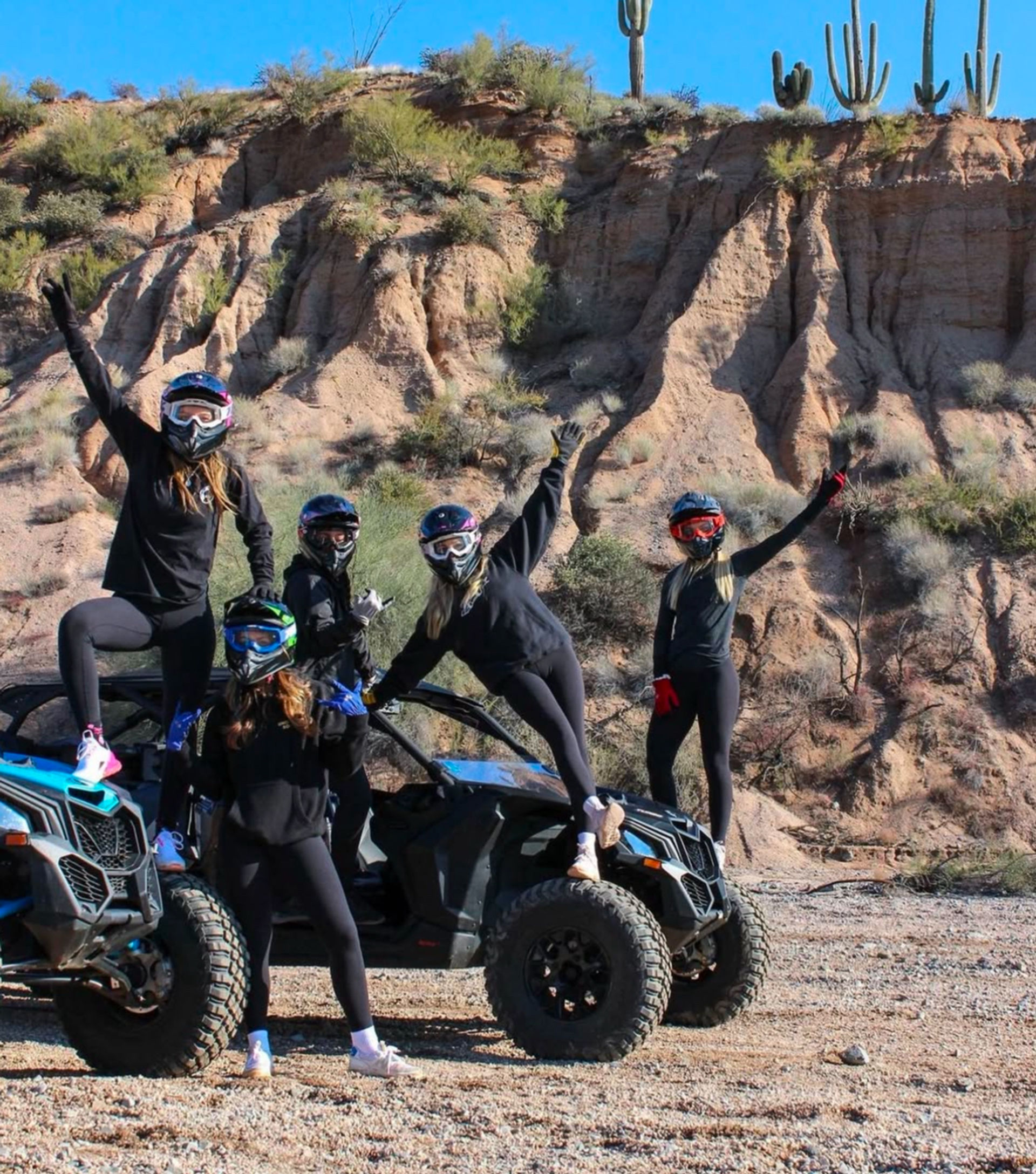 Five helmeted riders in black outfits posing on and beside two blue off-road UTVs in an Arizona desert, with saguaro cacti and red rock cliffs under a bright blue sky.