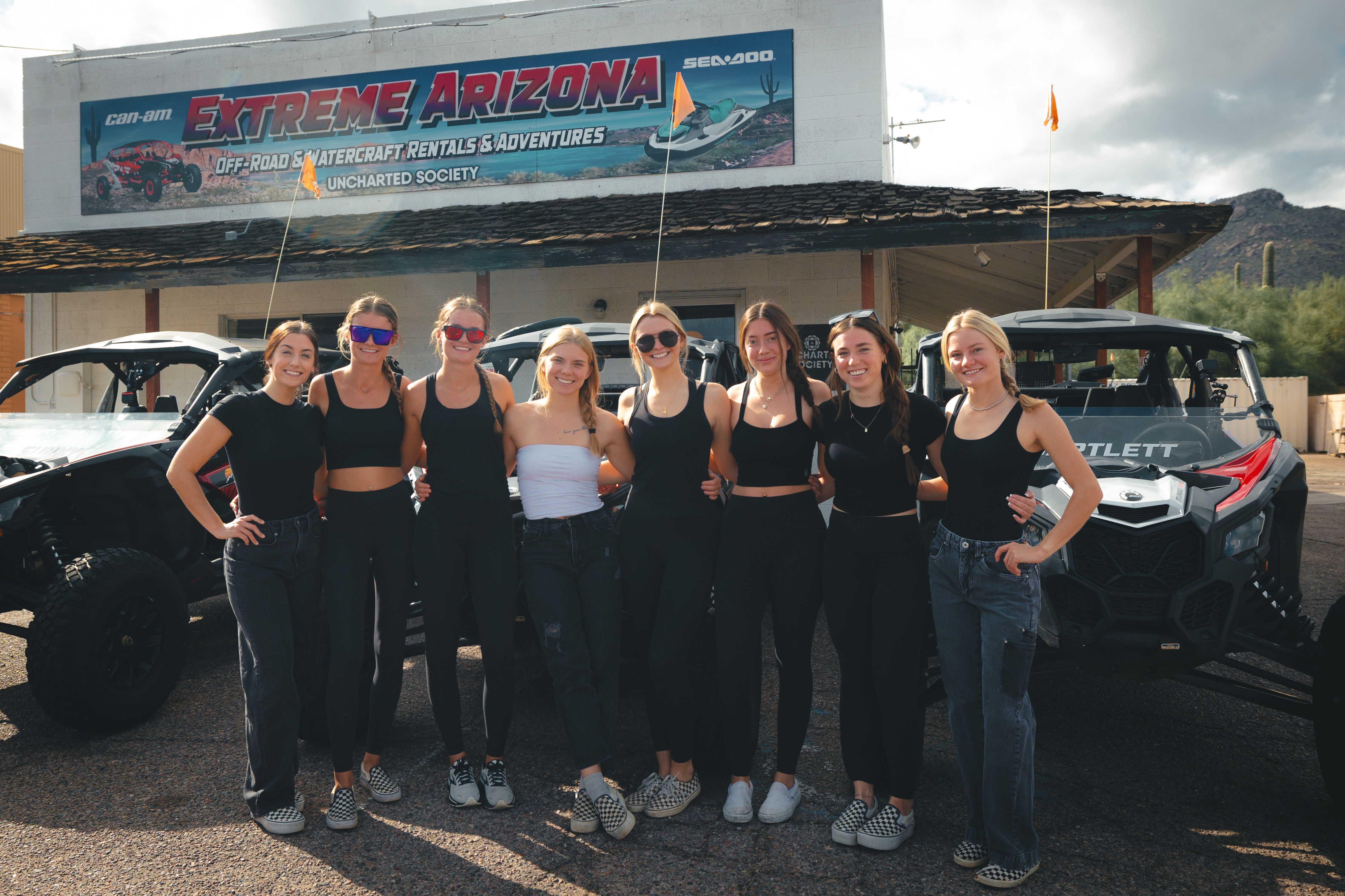 Eight smiling women in coordinated black outfits posing in front of off-road UTVs outside a rustic building in the Arizona desert with cacti and partly cloudy sky.