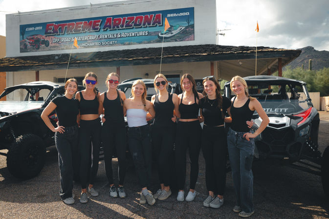 Eight smiling women in coordinated black outfits posing in front of off-road UTVs outside a rustic building in the Arizona desert with cacti and partly cloudy sky.