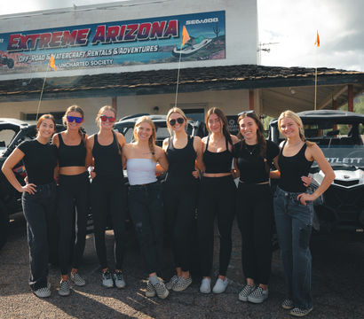 Eight smiling women in coordinated black outfits posing in front of off-road UTVs outside a rustic building in the Arizona desert with cacti and partly cloudy sky.