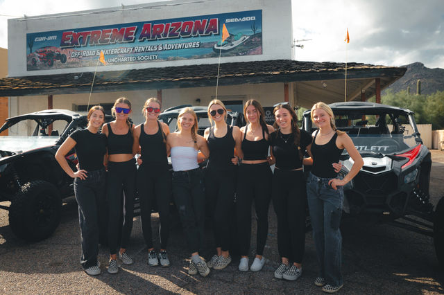 Eight smiling women in coordinated black outfits posing in front of off-road UTVs outside a rustic building in the Arizona desert with cacti and partly cloudy sky.