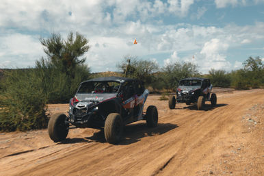 Two side-by-side off-road UTVs tearing across a sandy desert trail, kicking up dust past scrubby bushes under a bright, partly cloudy sky