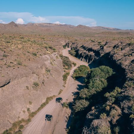 Aerial view of a dusty dirt trail winding through a rocky desert canyon with sparse scrub and two off-road vehicles heading toward distant mountains under a clear blue sky.