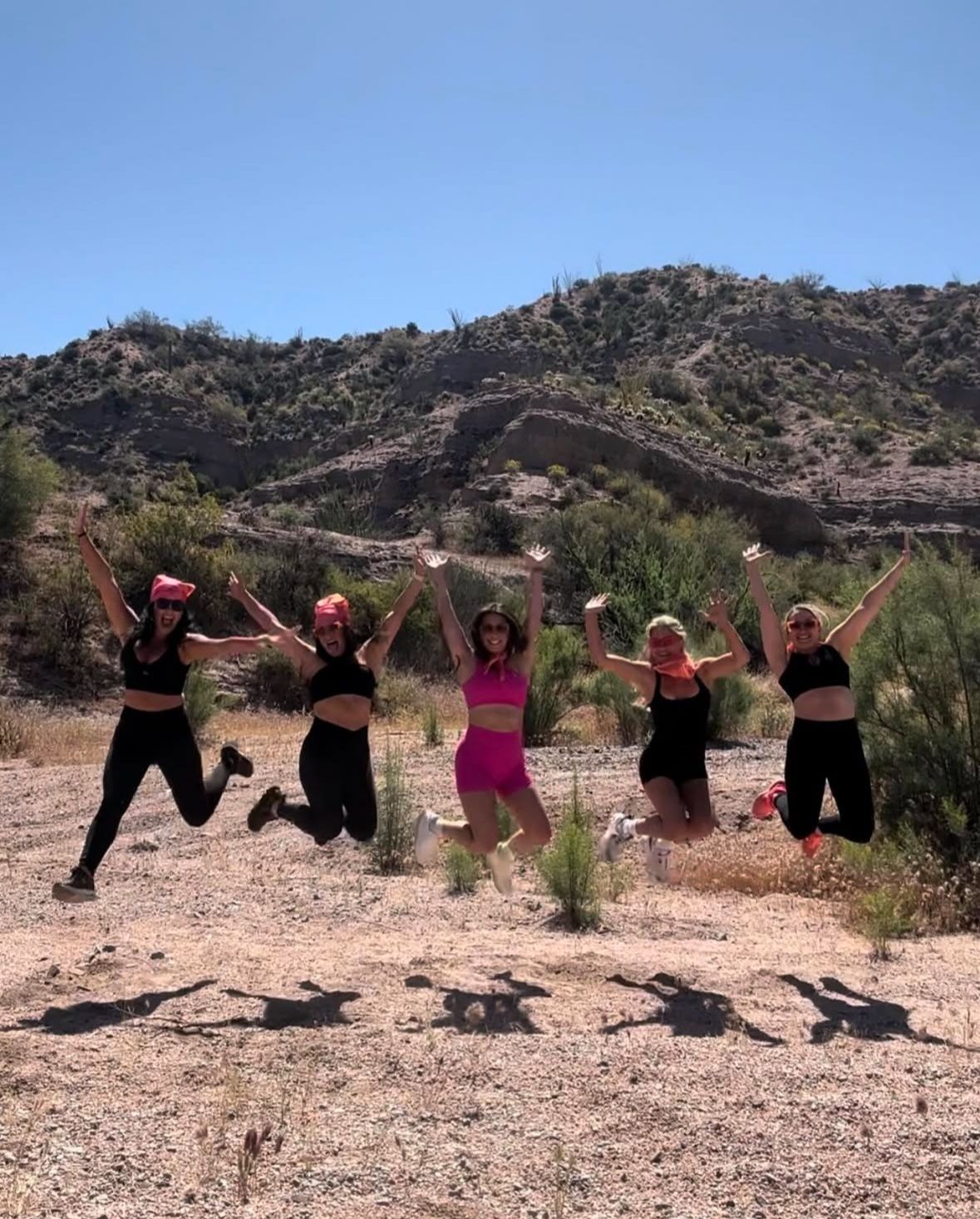 Five friends jumping midair on a sunlit desert trail with rocky hills and clear blue sky, wearing athletic outfits and bright bandanas