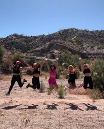 Five friends jumping midair on a sunlit desert trail with rocky hills and clear blue sky, wearing athletic outfits and bright bandanas