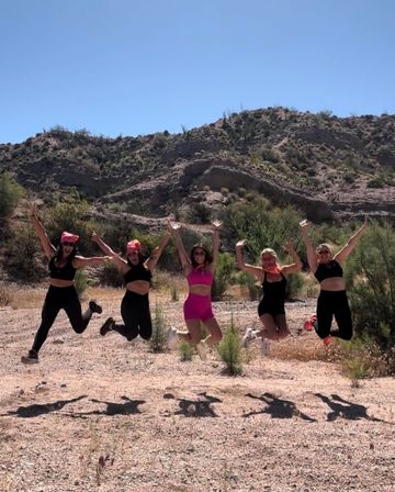 Five friends jumping midair on a sunlit desert trail with rocky hills and clear blue sky, wearing athletic outfits and bright bandanas
