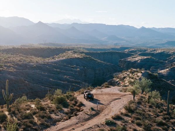 Off-road vehicle on a winding dirt trail above a sunlit Southwestern desert canyon dotted with saguaro cacti and layered mountain ridges.
