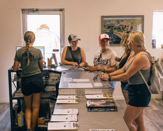 Group of women checking in at a metal-topped reception counter in an outdoor adventure outfitter office, staff at a computer, clipboards and brochures on the counter and a canyon landscape photo on the wall.