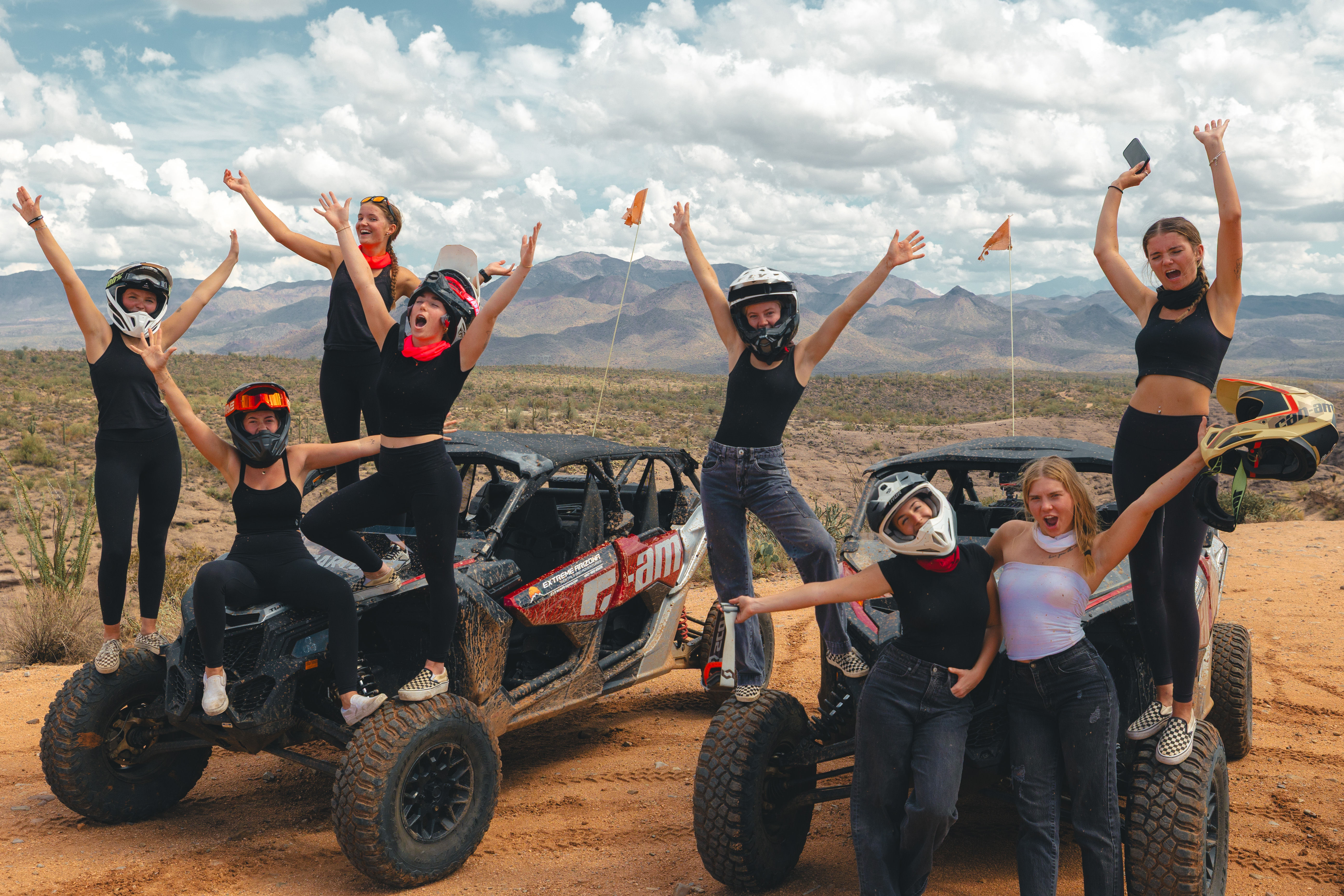 Energetic group of friends cheering and posing on two muddy off-road UTVs in a sunlit desert landscape with scrub, distant mountains, and dramatic cloudy sky — desert off-road adventure.
