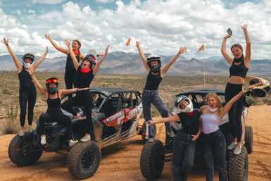 Energetic group of friends cheering and posing on two muddy off-road UTVs in a sunlit desert landscape with scrub, distant mountains, and dramatic cloudy sky — desert off-road adventure.