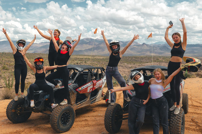 Energetic group of friends cheering and posing on two muddy off-road UTVs in a sunlit desert landscape with scrub, distant mountains, and dramatic cloudy sky — desert off-road adventure.