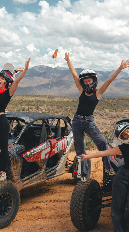 Energetic group of friends cheering and posing on two muddy off-road UTVs in a sunlit desert landscape with scrub, distant mountains, and dramatic cloudy sky — desert off-road adventure.
