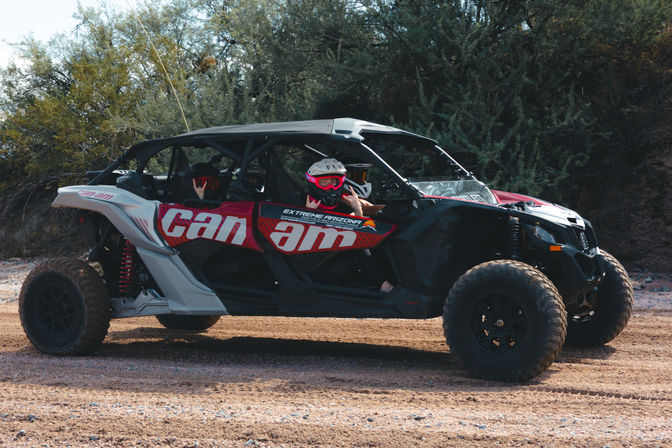 Two helmeted riders in a red-and-white off-road UTV side-by-side on a dusty desert trail, one flashing a peace sign amid scrubby bushes — outdoor off-road adventure.