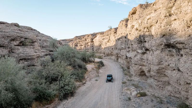 Aerial view of an off-road buggy on a winding dirt road through a sunlit arid canyon with towering rocky cliffs and sparse desert vegetation