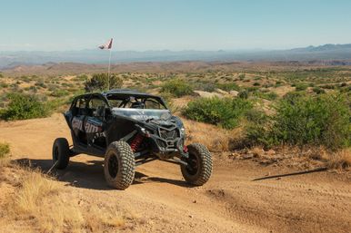 Rugged four-seat off-road UTV with a tall safety flag parked on a dusty desert trail, surrounded by scrub brush, rolling hills and distant mountain ranges under a bright blue sky