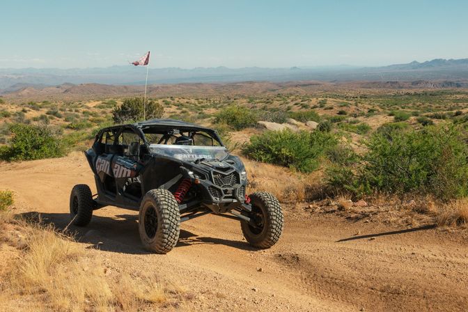 Rugged four-seat off-road UTV with a tall safety flag parked on a dusty desert trail, surrounded by scrub brush, rolling hills and distant mountain ranges under a bright blue sky