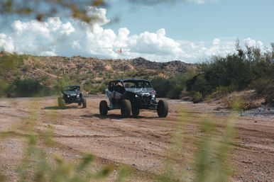 Two off-road side-by-side UTVs on a dusty desert trail with scrubby cacti and rocky hills, an orange safety flag, and dramatic cumulus clouds under a bright blue sky — ready for adventure.