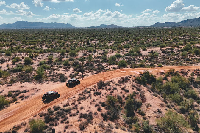 Two off-road buggies kicking up red dust on a winding dirt road through a saguaro-studded Southwestern desert with distant mountains and a blue sky