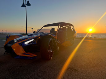 Orange-and-blue three-wheeled open sports car parked on an oceanfront road at sunset, golden sun and lens flare reflecting across the pavement and sea.