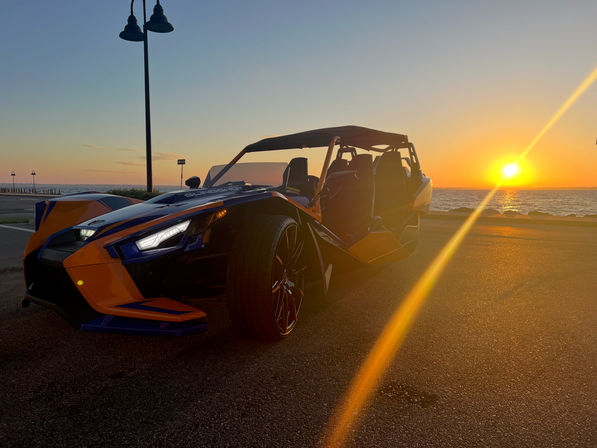 Orange-and-blue three-wheeled open sports car parked on an oceanfront road at sunset, golden sun and lens flare reflecting across the pavement and sea.